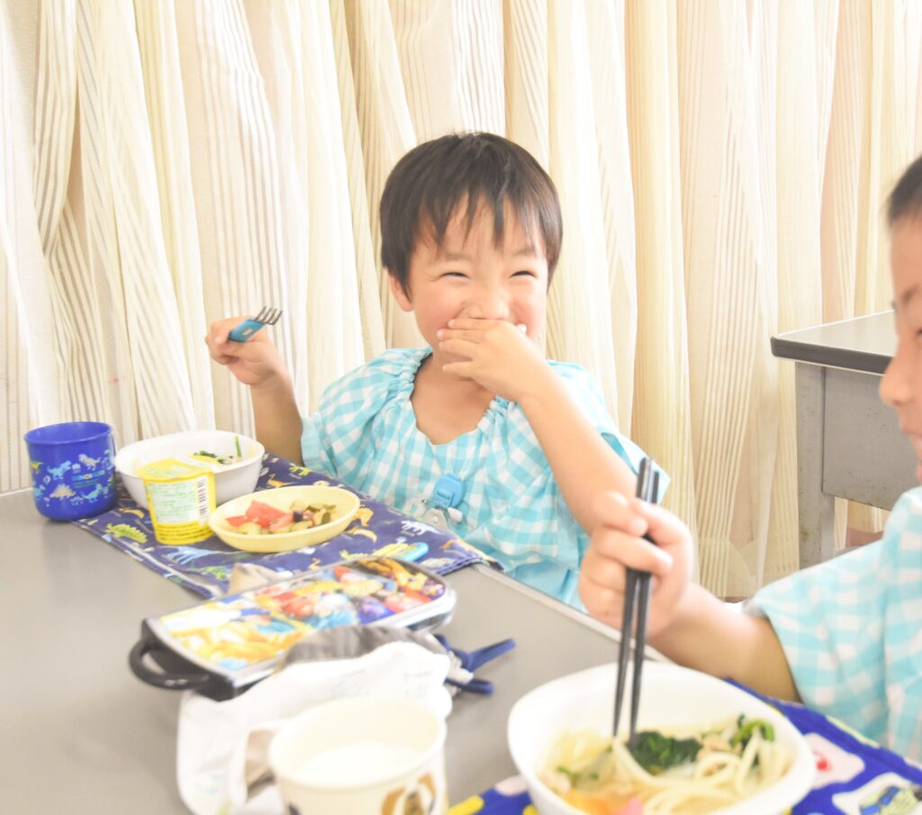 幼稚園で子どもたちが栄養バランスの良い給食を食べながら笑顔で過ごしている様子。みんなで楽しく食事を囲むシーン。
