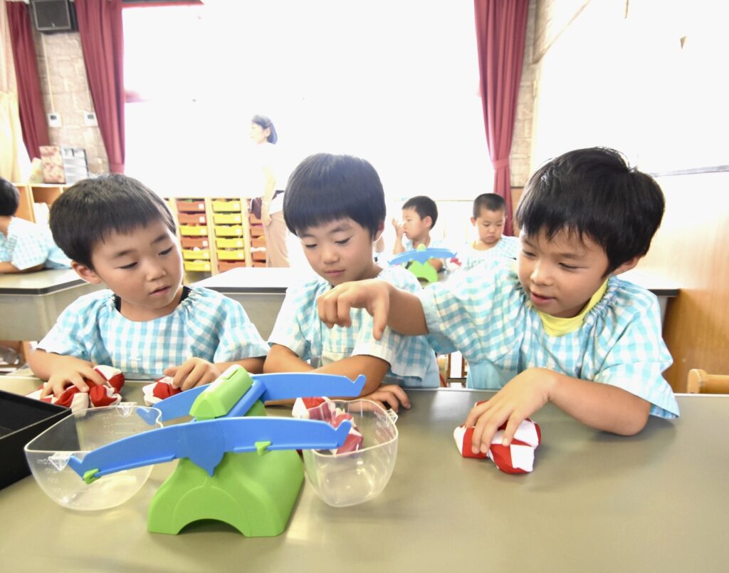 幼稚園で子どもたちが重さ比べの遊びを通してバランス感覚や数の概念を学んでいる様子。楽しそうに取り組んでいる。