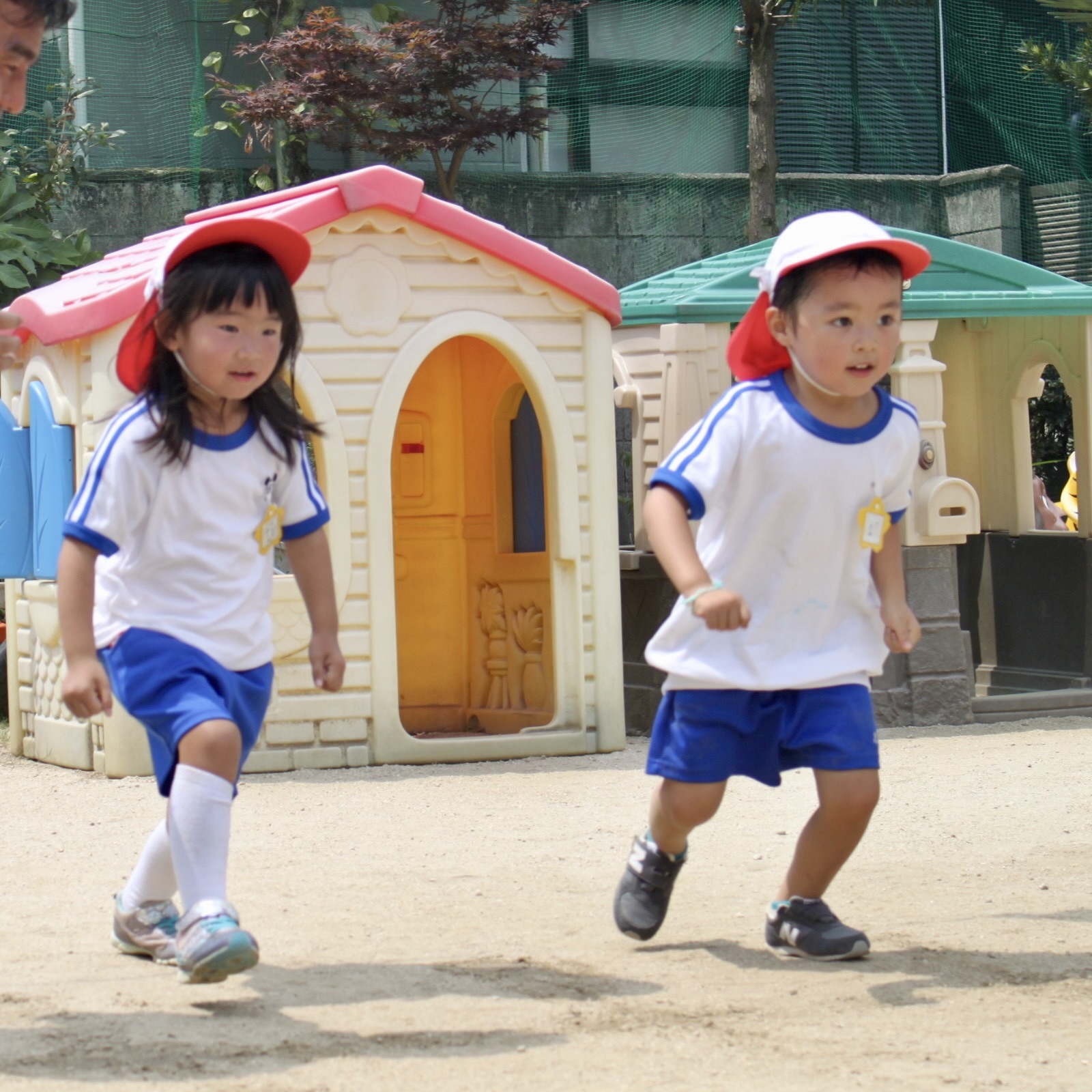 幼稚園の園庭で、白い帽子をかぶった子どもたちが元気よく走って遊んでいる様子。背景に小さな家の遊具がある。