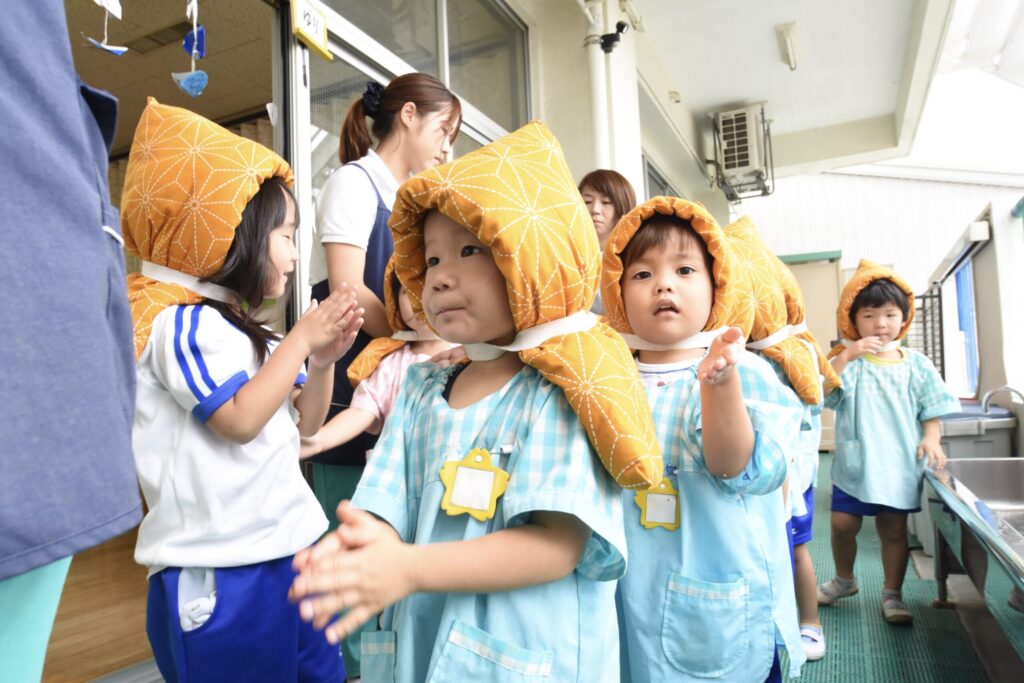 幼稚園で防災頭巾をかぶった子どもたちが避難訓練をしている様子。先生と一緒に外へ出て並んでいる場面。