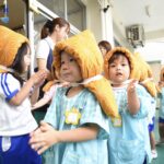 幼稚園で防災頭巾をかぶった子どもたちが避難訓練をしている様子。先生と一緒に外へ出て並んでいる場面。