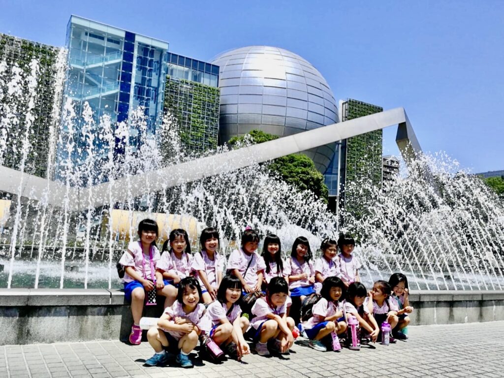 噴水の前でポーズをとる幼稚園児たち。科学館の建物を背景にした集合写真で、青空の下で楽しそうに笑っている。