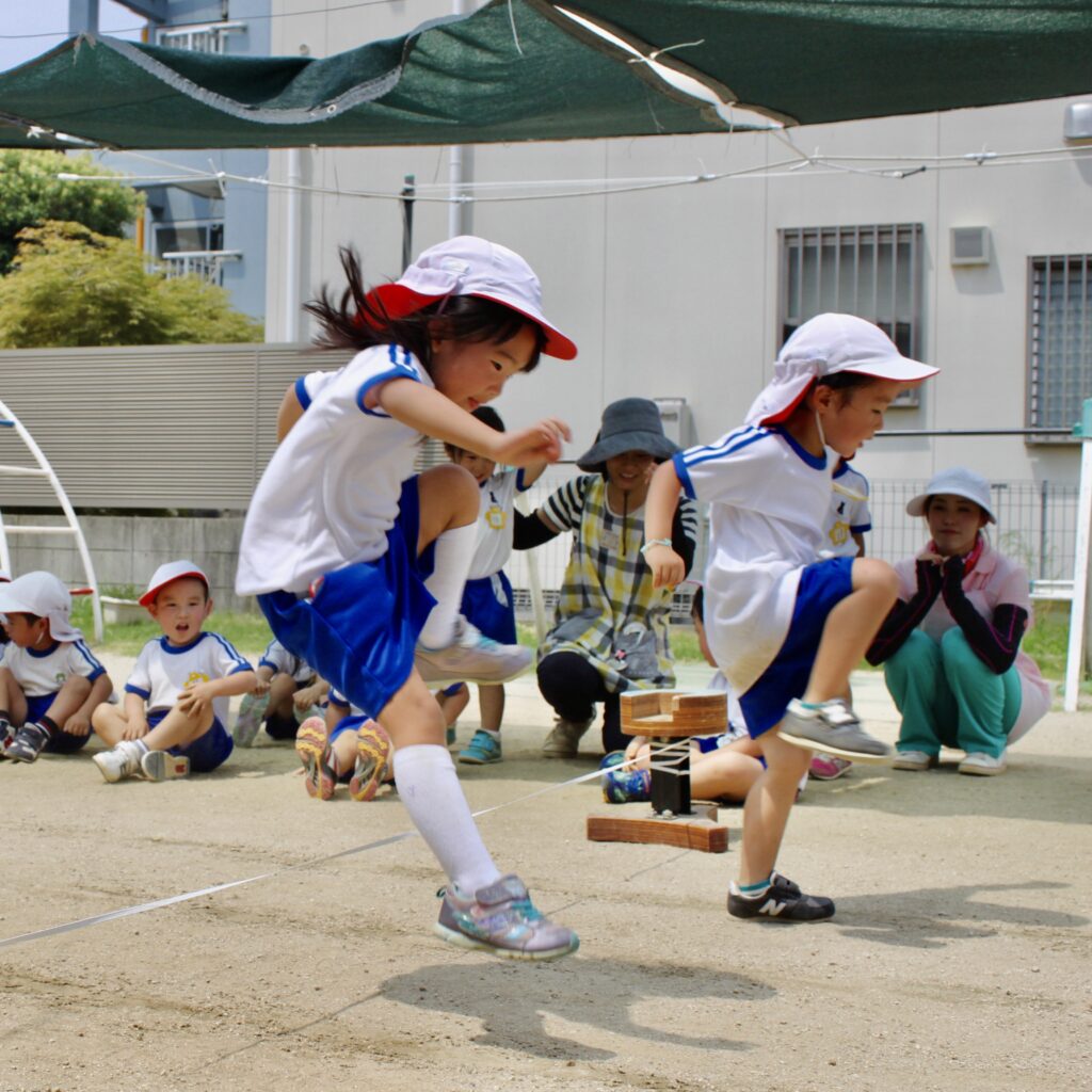 幼稚園の園庭で、子どもたちがジャンプする遊びをしている様子。元気いっぱいに体を動かしている場面。