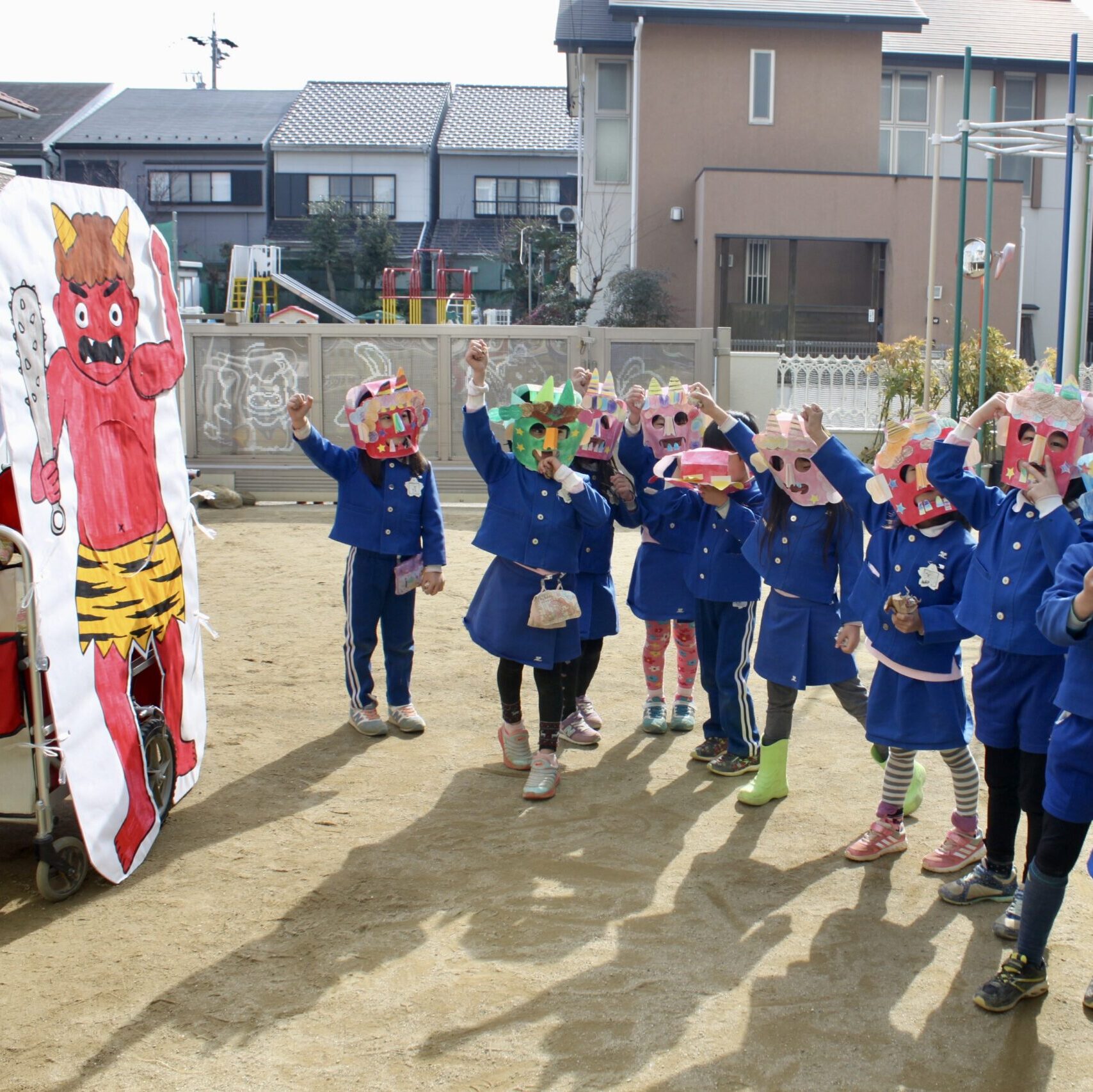 幼稚園の園庭で、鬼のお面をかぶった子どもたちが豆まきをして節分を楽しんでいる様子。背景に赤鬼の絵が飾られている。